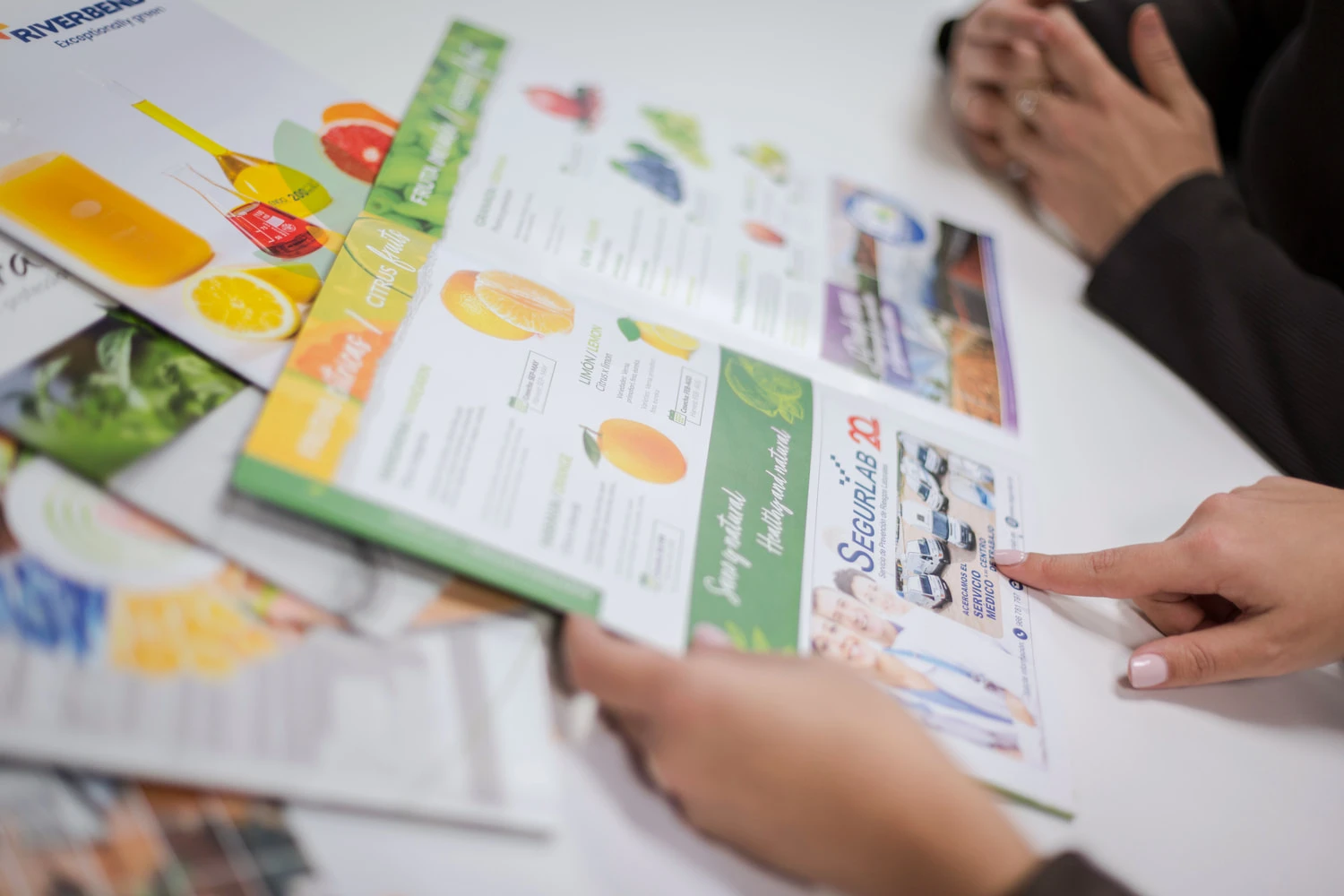 A bunch of brochures on a table and a woman's hands pointing in a brochure at JS Comunicación.